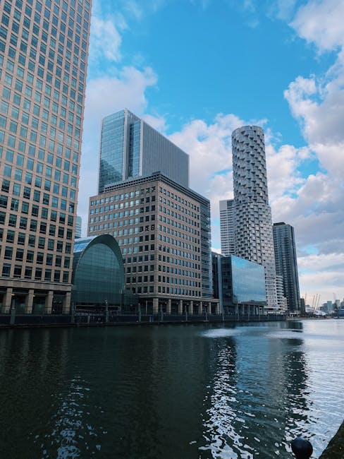 Black and white aerial view of a modern Canary Wharf office building with a curved façade, featuring multiple rows of windows on each floor. Surrounding the central building are other high-rise offic