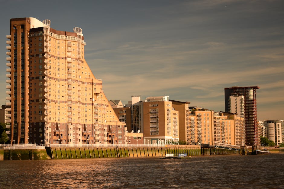 A wide view of a modern cityscape along a river, featuring tall buildings with glass and steel facades on both sides. The buildings show a mix of architectural styles, with some having reflective surf